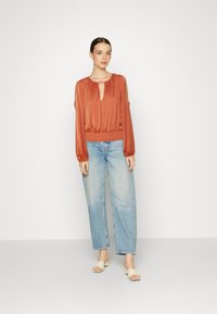 Blusa de cor ferrugem com decote em forma de chave e mangas longas, combinada com jeans de perna larga de cor azul claro e sandálias de salto bloco em tom creme.