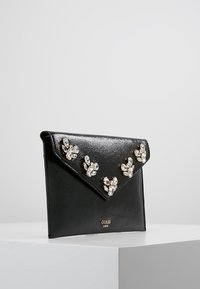 Svart fusk läder clutch med en spetsig klaff, prydd med kristalliknande detaljer och en gyllene logotyp längst ner, slät struktur.