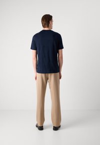 Jūrinio mėlynumo trumpas rankovėmis marškinėliai su subtiliu tekstūriniu raštu, derinami su smėlio spalvos plačiomis kelnėmis ir juodais batais, matoma iš nugaros.