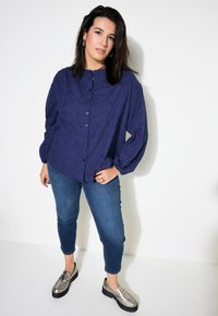Camicia azzurra con bottoni e motivo testurizzato, abbinata a jeans blu aderenti e scarpe slip-on metalliche. Sfondo bianco pulito.