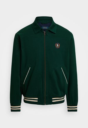 Žalia zip-up striukė su smailiu apykakle, baltais juostelėmis ir logotipo pleistru kairėje krūtinės pusėje. Turi dvi priekinės kišenes.