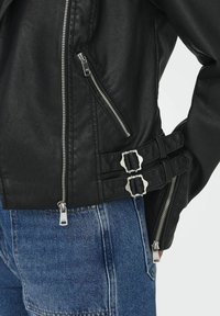 Veste en simili cuir noir avec fermetures éclair argentées, poches latérales et détails de boucle à la taille. Texture lisse et design ajusté.