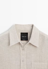 Light beige textured button-up shirt with pointed collar, two buttons visible, chest pocket, and black label reading "Massimo Dutti."