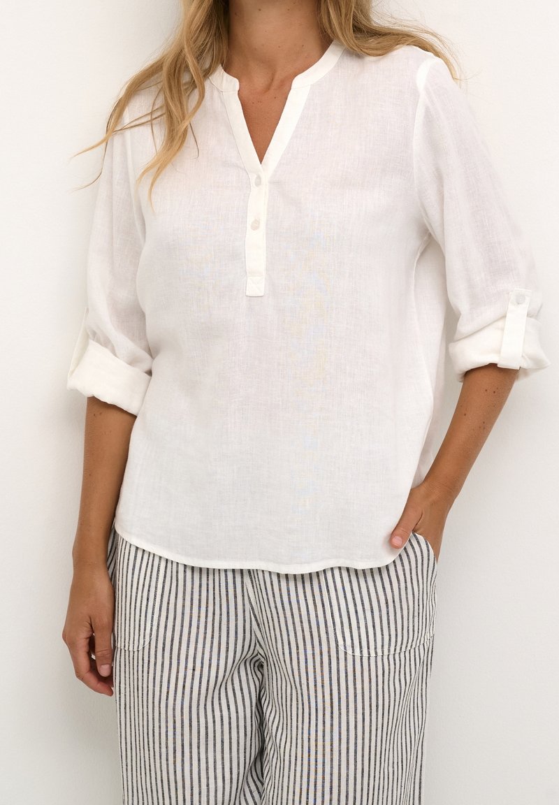 Femme portant une chemise blanche à manches longues avec les poignets retroussés et un pantalon à rayures verticales noires et blanches, debout avec une main dans la poche.