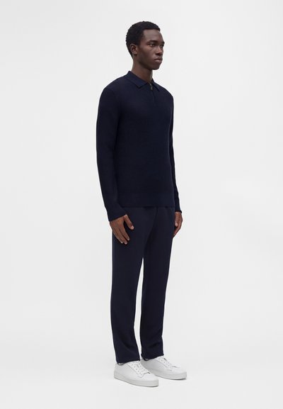 Homme debout avec un pull en maille bleu marine, un pantalon bleu marine assorti et des baskets blanches sur un fond clair uni.
