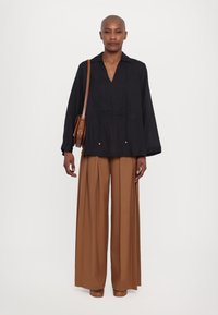 Blusa nera con scollo a V, maniche ampie e piccoli accenti metallici; abbinata a pantaloni marroni a vita alta e a gamba larga; borsa a spalla marrone.