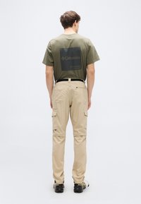 Ένα άτομο φοράει χακί παντελόνι cargo και πράσινη ελιά κοντομάνικη μπλούζα με μεγάλο μαύρο λογότυπο στην πλάτη. Φαίνονται μαύρα παπούτσια.