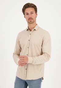 Camisa beige de manga larga con cuello, cierre frontal de botones y un logo sutil. Presenta un dobladillo recto y una textura de tela suave.