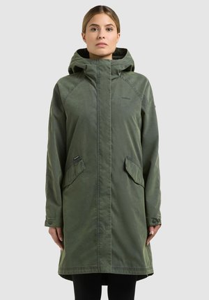 Woman wearing a long, olive green hooded coat with flap pockets, standing against a plain light background.