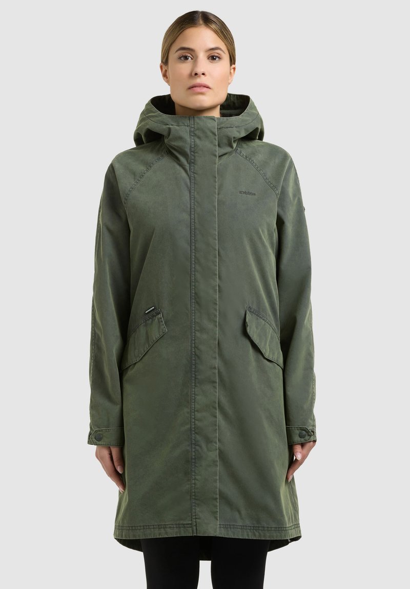 Femme portant un long manteau à capuche vert olive avec des poches à rabat, debout devant un fond clair uni.