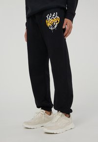Pantaloni sportivi neri con una grafica "Feel Happy" in bianco e giallo, dotati di una vita elasticizzata e polsini affusolati. Indossati con sneakers chiare.