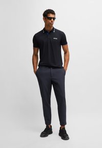 Crna polo majica s plavim obrubima i bijelim logotipom, u kombinaciji s tamnim hlačama i crnim tenisicama. Model nosi sunčane naočale.