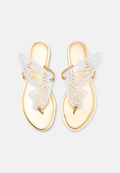 Sandales plates dorées avec des brides en forme de papillon blanches et des anneaux pour les orteils sur un fond clair.