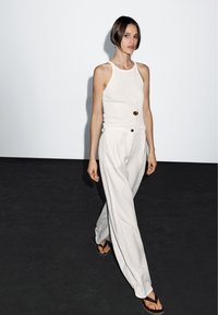 Witte tanktop en lichtbeige wijde broek met knoopdetail. Schoeisel bestaat uit bruine flip-flops. Eenvoudige zilveren ketting met hanger.