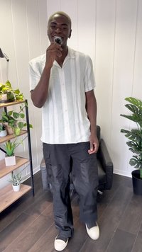 Licht gestreepte kortekolenhemd in wit en groen, gecombineerd met zwarte cargo-broeken en lichtgekleurde schoenen, met kamerplanten op de achtergrond.