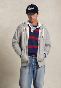 Gray zip-up hoodie with white drawstrings, layered over a navy and red striped polo shirt, paired with light blue denim jeans.