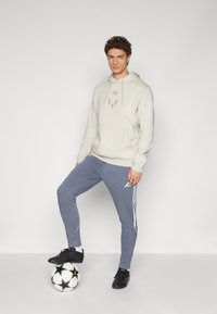 Beige Kapuzenpulli mit einer Vorderstasche und Logo, kombiniert mit grauen Jogginghosen mit weißen Streifen. Schwarze Fußballschuhe auf einem gemusterten Ball.
