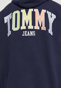 Jūras spēku kapuce ar daudzkrāsainu izšuvumu "TOMMY" un baltu "JEANS", izceļot mīkstu tekstūru un brīvu piegriezumu.