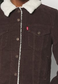 Giacca in corduroy marrone con colletto in pile bianco, bottoni a pressione in metallo, tasche a pattina sul petto e una piccola etichetta rossa del marchio sul lato sinistro.