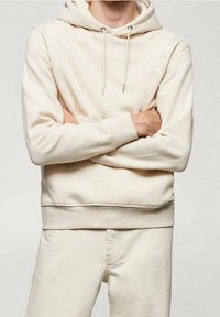 Personne portant un sweat à capuche de couleur crème avec des cordons et un pantalon assorti, les bras croisés devant la poitrine.