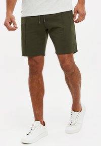 Pantalones cortos de algodón de mezcla verde oscuro con cintura elástica, cordón y bolsillos delanteros, combinados con zapatillas blancas. Textura suave.