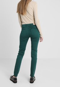 Pantalones verdes de mezcla de algodón con corte ajustado, cinco bolsillos y una etiqueta en la cintura. La tela tiene una textura suave y un ligero brillo.