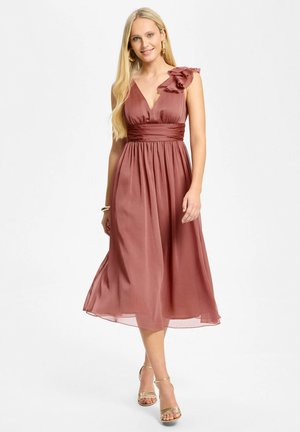 Femme portant une robe midi rose poudré sans manches avec une épaule volantée et des sandales à talons hauts dorées, souriante sur fond blanc.
