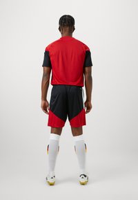 Κόκκινη και μαύρη αθλητική φανέλα με πλέγμα, συνδυασμένη με μαύρες βερμούδες. Λευκές κάλτσες μέχρι το γόνατο με χρωματιστές λεπτομέρειες. Εμφανίζεται η πίσω όψη.