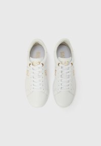 Fehér sportcipők sima bőr textúrával, arany EA7 márkajelzéssel az oldalukon, fehér fűzőkkel és alacsony szárú dizájnnal.