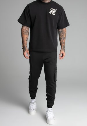 Man wearing black short-sleeve t-shirt with white logo, black cargo joggers, white sneakers, and visible arm tattoos against gray background.