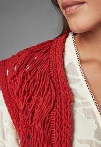Gilet rosso texture con un lungo design frangiato, indossato sopra un capo bianco con un motivo floreale e scollatura a scallop.