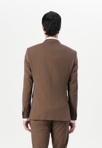 Jachetă de costum maro cu o textură netedă, croi ajustat și ventilație unică. Dispune de mâneci lungi și detalii de cusătură subtile, asociată cu pantaloni asortați.