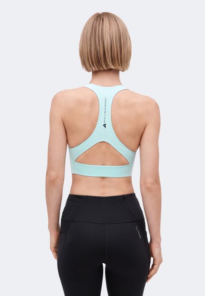Frau mit kurzen blonden Haaren, die einen hellblauen Racerback-Sport-BH und schwarze, hoch taillierte Leggings trägt, mit dem Rücken zu einem schlichten Hintergrund.