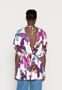Donna che indossa una blusa a fantasia colorata con scollatura a V profonda e colletto annodato, abbinata a jeans blu, in piedi contro uno sfondo semplice.