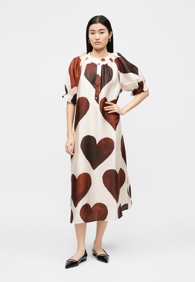 Γυναίκα που φοράει λευκό midi φόρεμα με μεγάλα σκούρα κόκκινα σχέδια καρδιών, φουσκωτά μανίκια, κουμπωτό ντεκολτέ και μαύρες μυτερές μπαλαρίνες σε απλό φόντο.