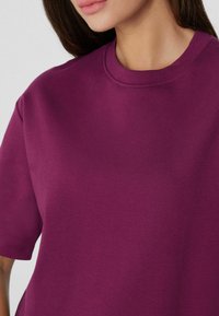 Camiseta oversized de color púrpura oscuro con cuello redondo y mangas cortas, hecha de una tela suave con una textura delicada. Detalles de diseño minimalista.