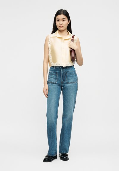 Γυναίκα με αμάνικο μπεζ μπλουζάκι με δαντελωτό τελείωμα, τζιν ψηλόμεσο μπλε με μπροστινές τσέπες, μαύρα μοκασίνια και κρατά καφέ τσάντα ώμου.