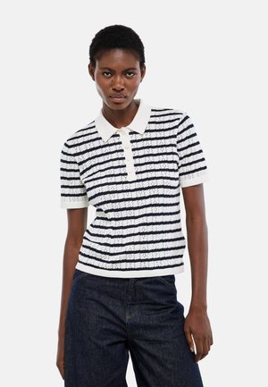 Femme portant un polo en maille à manches courtes rayé blanc et marine, associé à un jean taille haute bleu foncé, debout devant un fond uni.