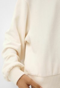 Kremowy długi rękaw sweatshirt z prążkowanymi mankietami, noszony przez osobę z jedną ręką blisko dolnego brzegu.
