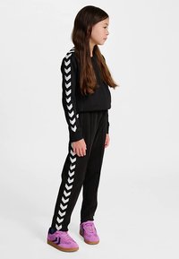 Czarny dres z białymi paskami w kształcie karo na rękawach i bokach, wykonany z gładkiej tkaniny. W połączeniu z fioletowymi trampkami i prostymi włosami.