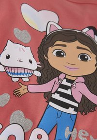 Rosa T-Shirt mit einem Cartoon-Mädchen mit lockigem Haar, das ein gestreiftes Shirt und eine Kapuzenjacke trägt und ein niedliches Tier-Cupcake-Charakter hält, mit glitzernden Herzakzenten.