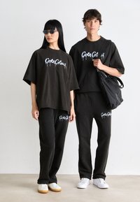 Twee modellen dragen bijpassende zwarte trainingssets met witte grafische tekst, waarbij één een zwarte tas vasthoudt, staand tegen een effen lichte achtergrond.