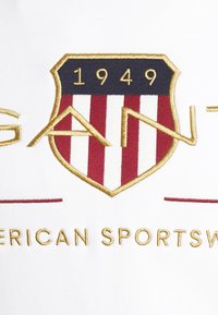 Weißer Stoff mit einem gestickten Wappenlogo, das rote und blaue Streifen, goldene Details und den Text "GANT" zeigt, mit "1949" darüber und "AMERICAN SPORTSWEAR" darunter.