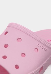 Κοντινή λήψη ενός ανοιχτόχρωμου ροζ Crocs clog που δείχνει οπές αερισμού και υφή στην πάνω λουρίδα με ανάγλυφο το όνομα της μάρκας.