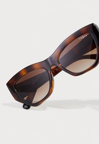 Ochelari de soare supradimensionați în stil cat-eye, cu un model de broască țestoasă, lentile degradate de culoare brună și o finisare lucioasă. Prezentă cuvântul "vogue" pe rame.