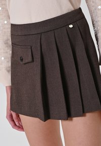 Gonna mini a pieghe color marrone con tasca a flap frontale con bottoni, indossata con un top leggero a maniche lunghe con dettagli in paillettes sulle maniche.