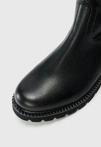 Botte de cheville en cuir noir avec une finition lisse, bout arrondi et semelle en caoutchouc texturée comportant des accents pointus le long du bord.
