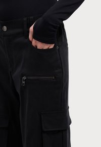 Handen i fickan på svarta cargobyxor med dragkedje- och ficklock, bärs tillsammans med en svart långärmad topp.