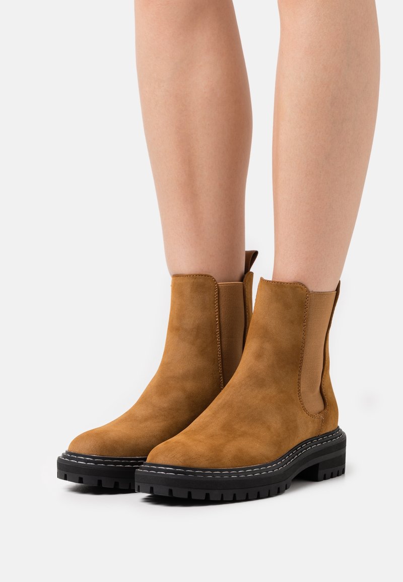 Personne portant des bottes mi-mollet en daim marron avec des semelles épaisses noires et des panneaux latéraux extensibles, debout sur une surface blanche.