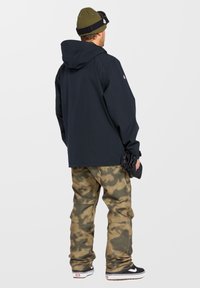 Schwarze Kapuzenjacke mit lockerer Passform, kombiniert mit camouflagefarbigen Cargo-Hosen. Getragen mit schwarz-weißen Sneakers und einer grünen Mütze.